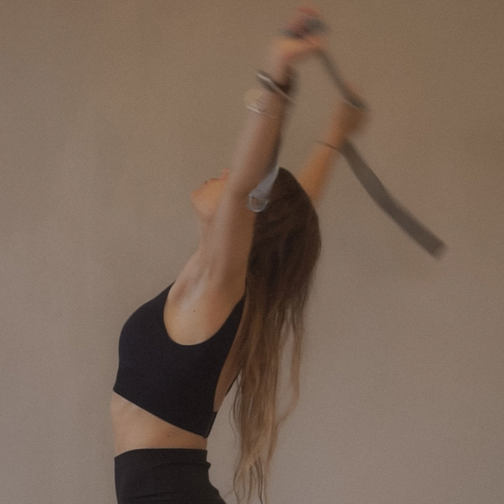 Woman in black sportswear stretching with arms raised and head tilted back against a plain wall.