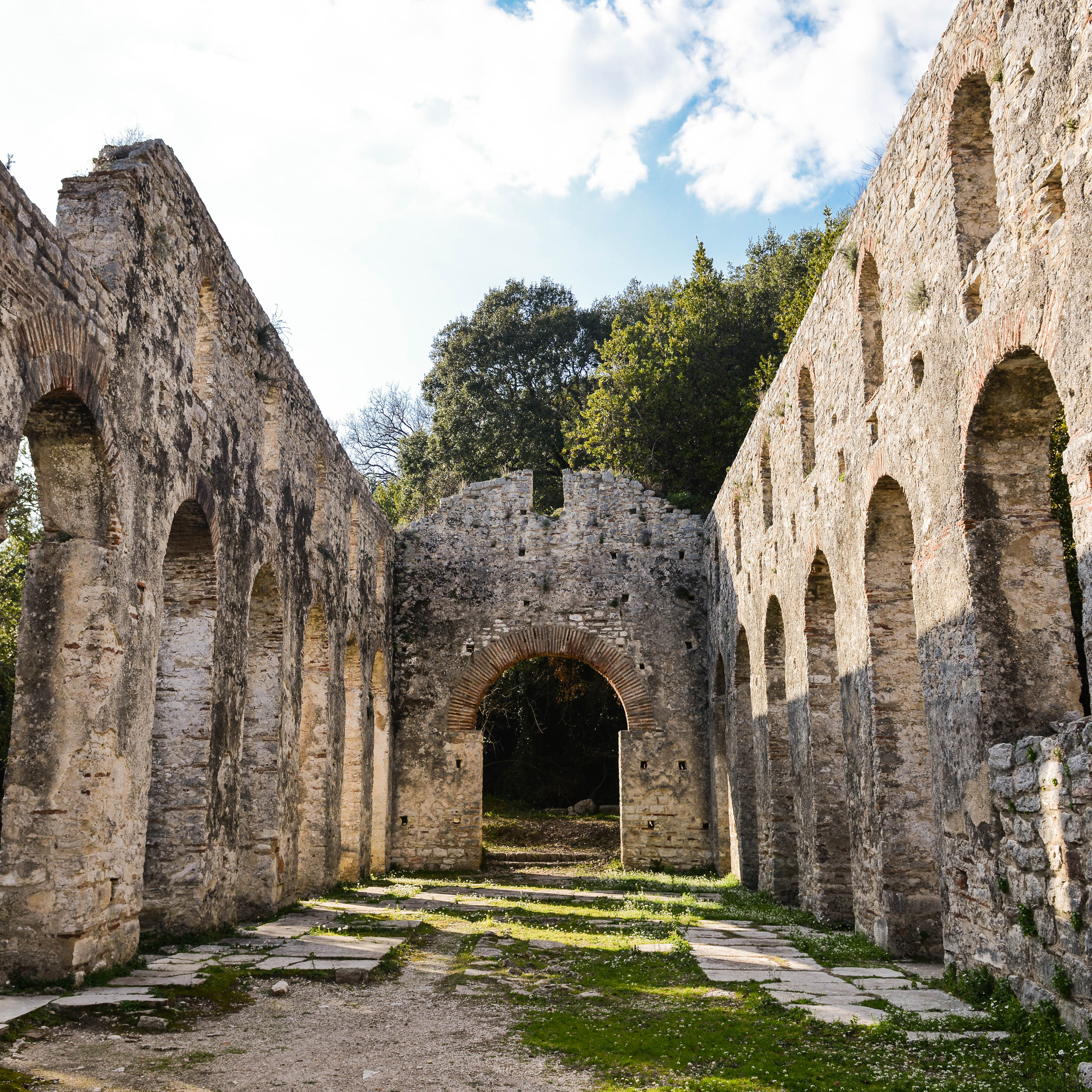 Ruins of an ancient stone structure with tall arched walls and an arched entrance, surrounded by greenery and trees.