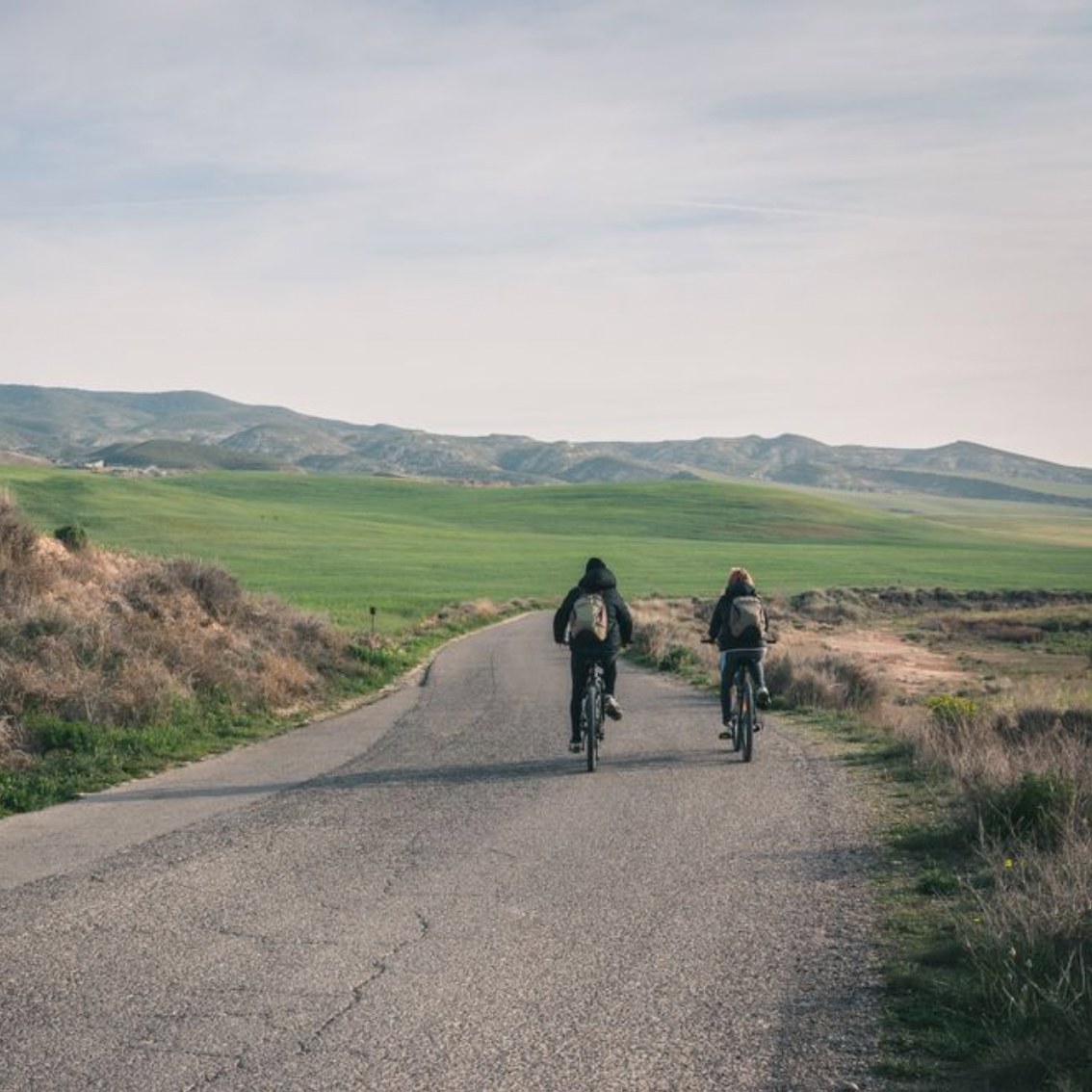 Two people riding bicycles on a paved road through a green and brown landscape with hills in the background.