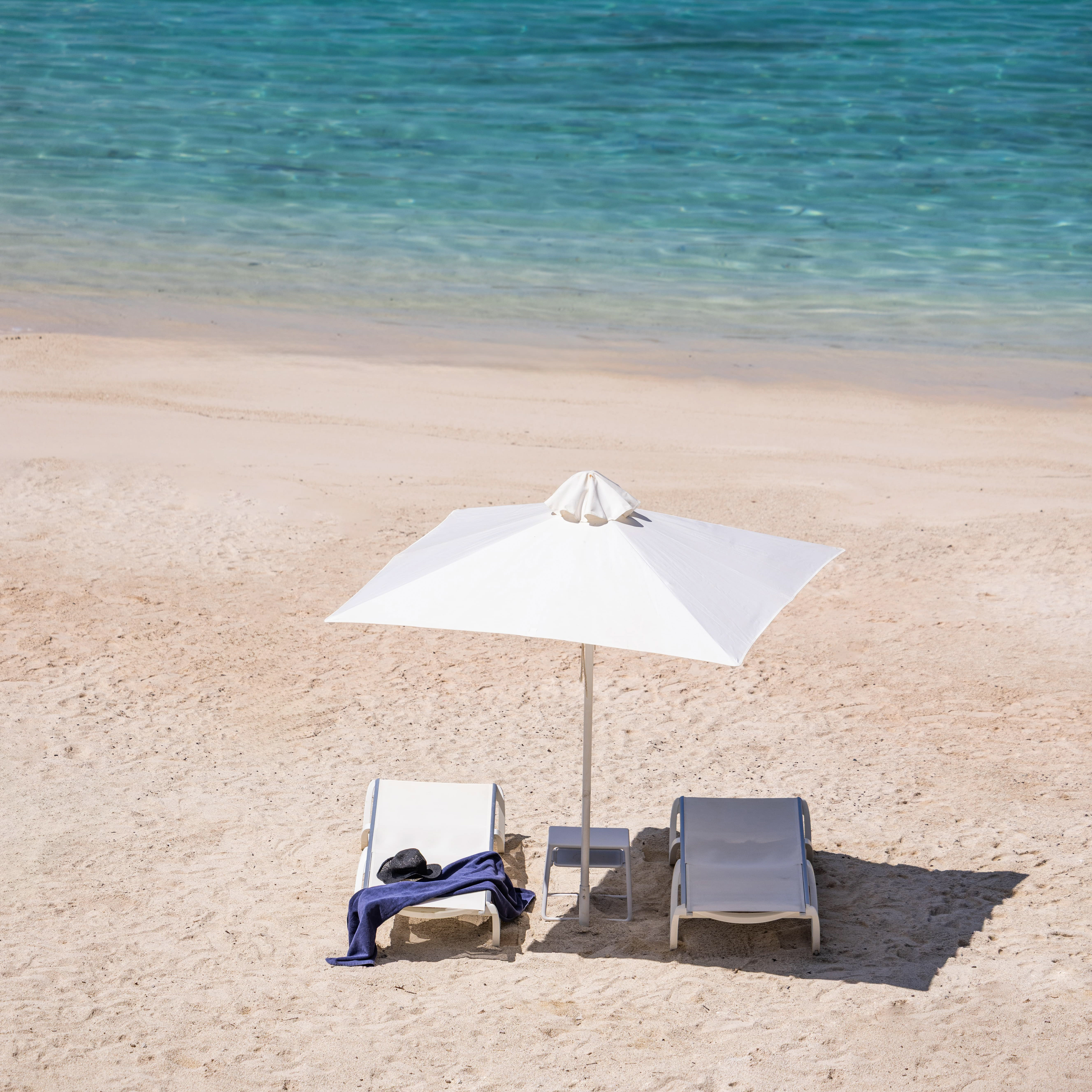 Two empty beach lounge chairs under a white umbrella on sandy shore with blue towel and hat on left chair.