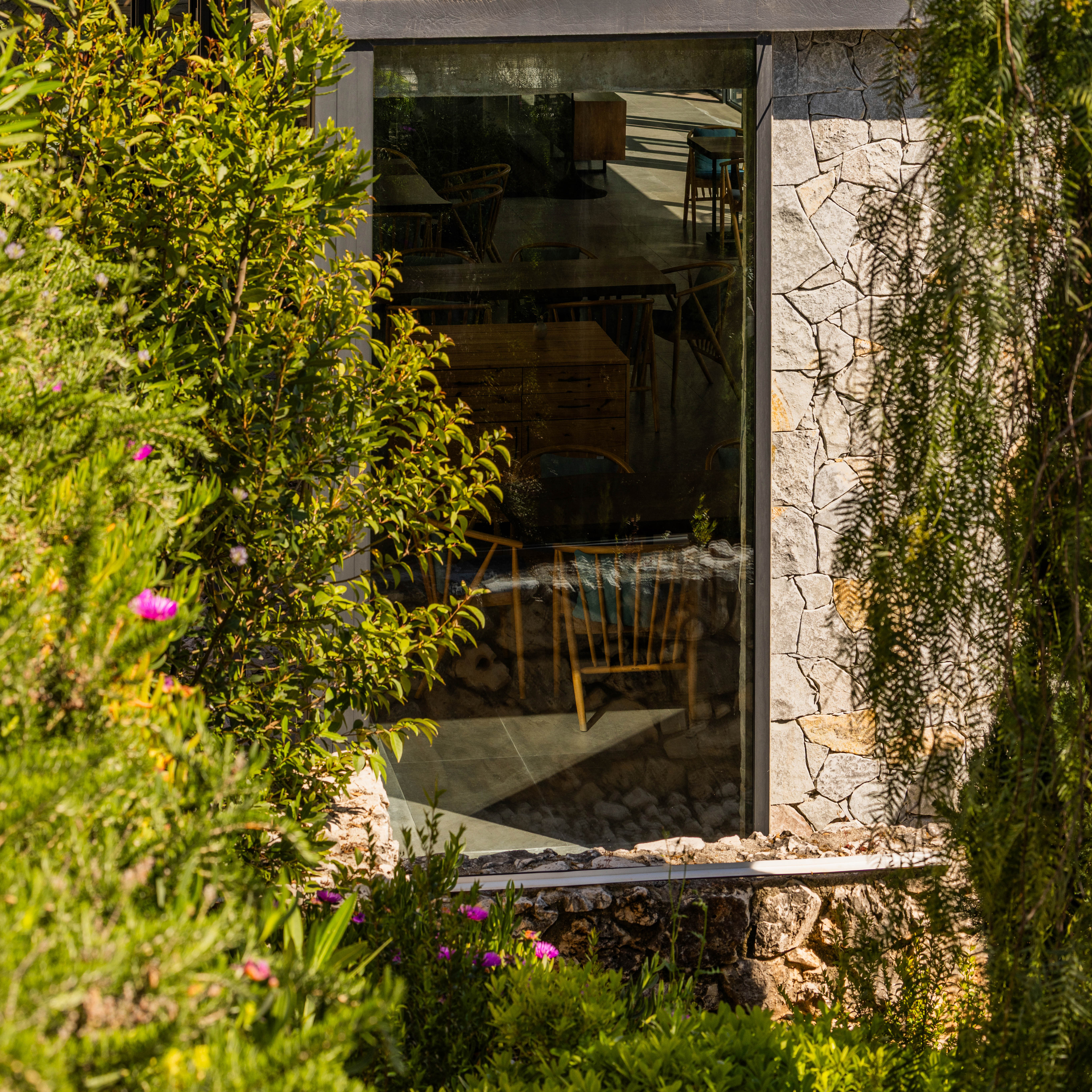 Large window with reflection of interior wooden chairs and tables, surrounded by green plants and stone wall exterior.