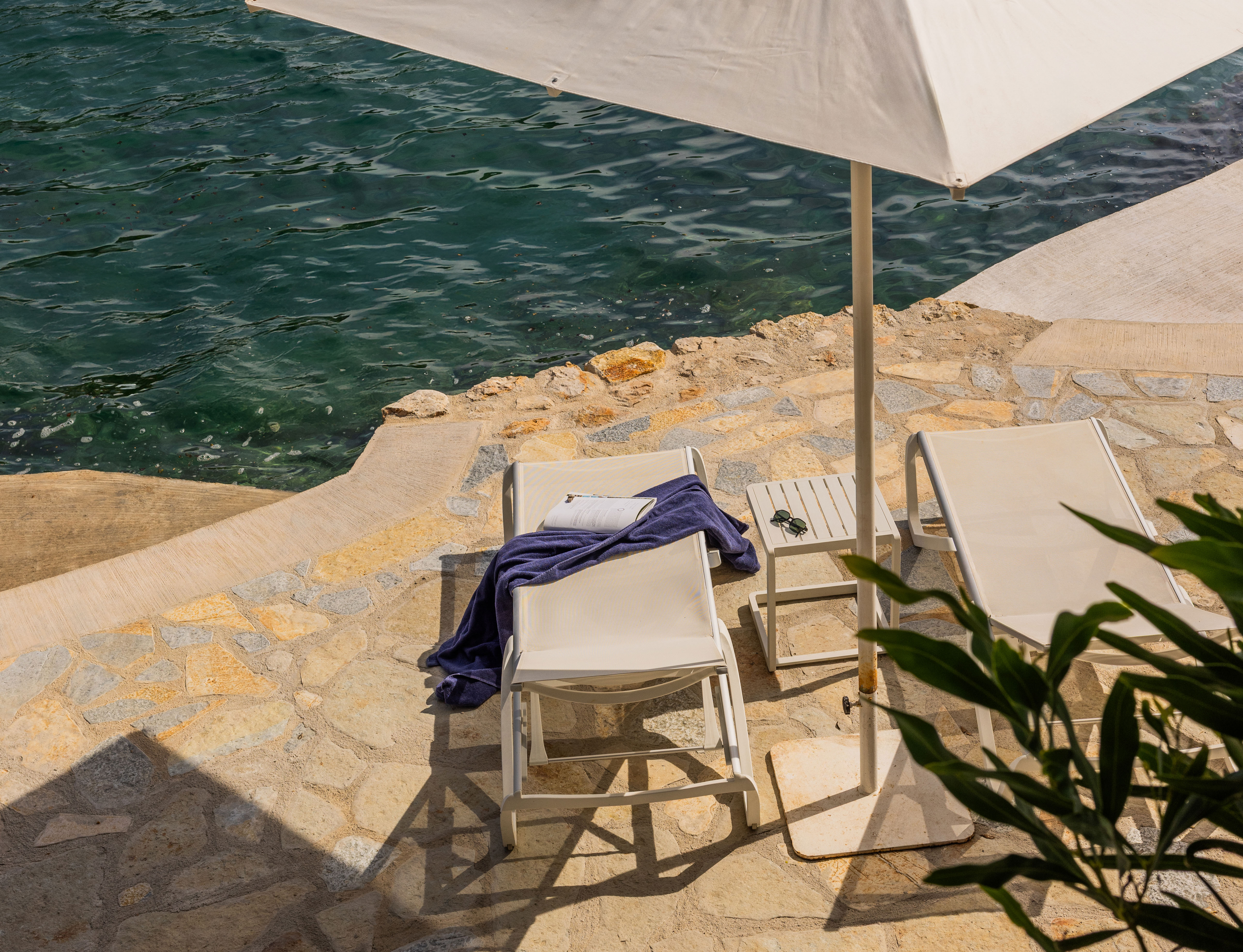 Two white lounge chairs with a blue towel and a book on one, and sunglasses on a small table between them by clear water under a large white umbrella.