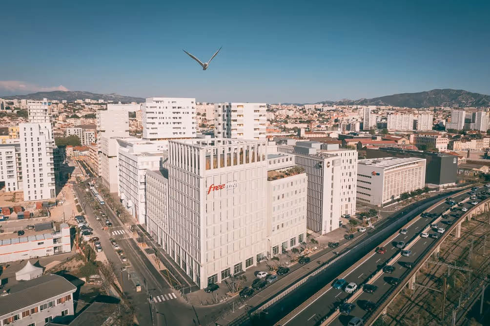 Aerial view of a modern urban cityscape with white high-rise buildings, including the 'Free Pro' headquarters, adjacent to a busy multi-lane highway and train tracks, with a bird flying overhead and mountains in the distance.