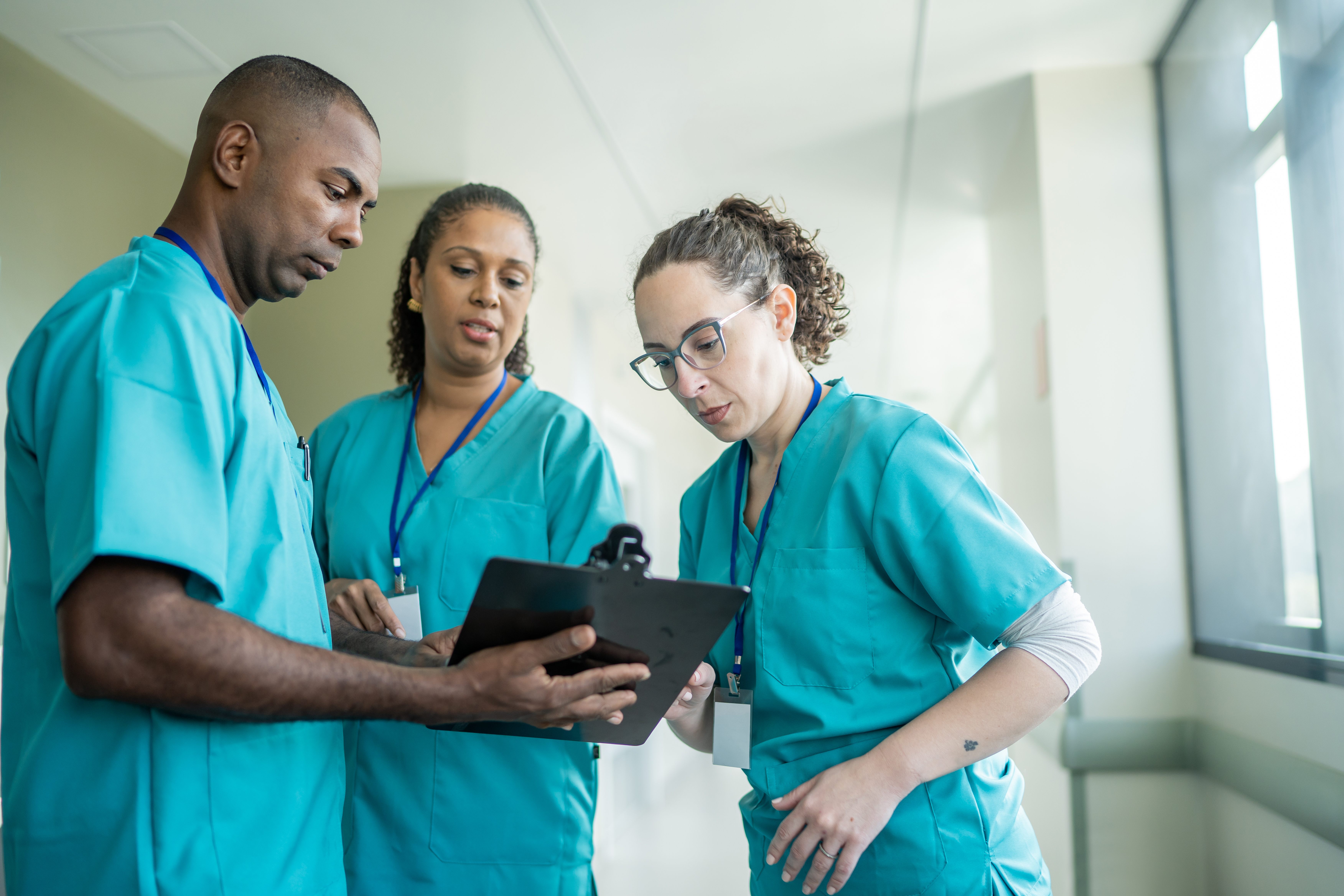 Three healthcare professionals in scrubs reviewing a clipboard together in a brightly lit hospital hallway, engaged in a focused discussion.