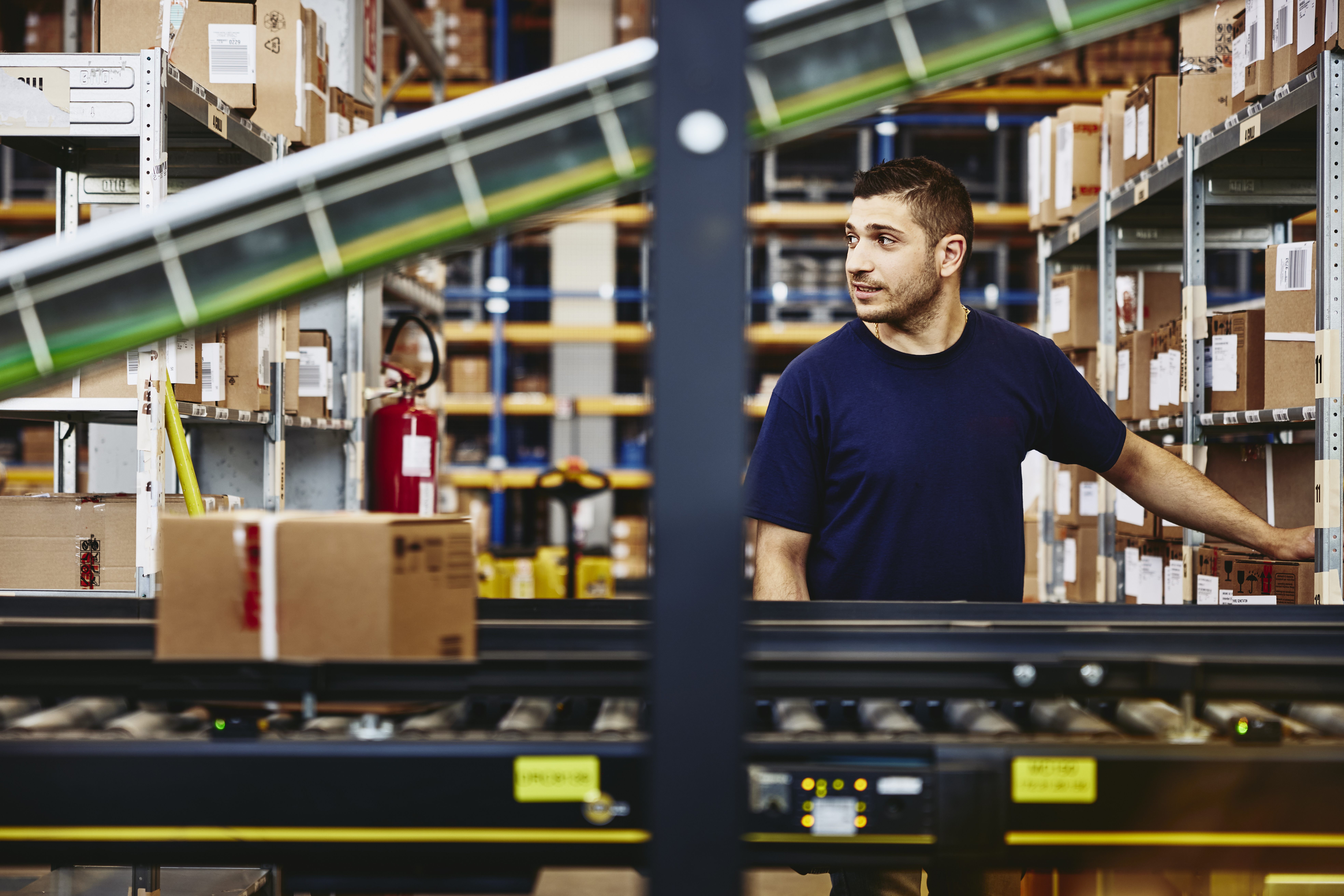 Warehouse worker organizing inventory on shelves near a conveyor belt, with cardboard boxes and packages visible throughout the industrial storage area.