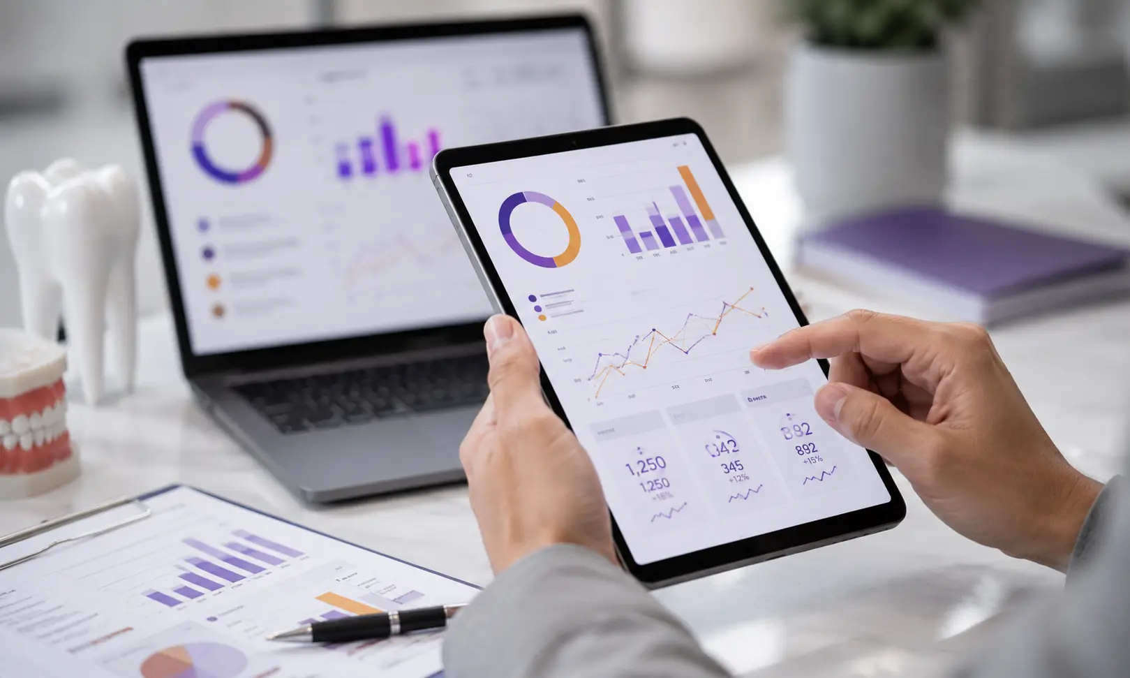 Person using tablet displaying graphs and charts with laptop and dental models on desk.