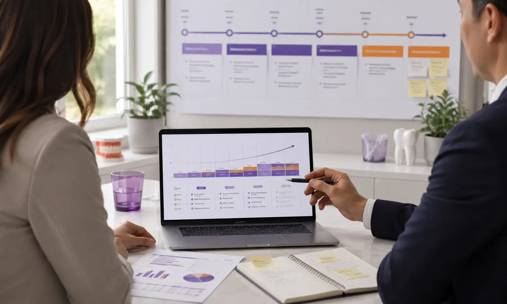 Two professionals review data charts and graphs on a laptop and documents at a desk.