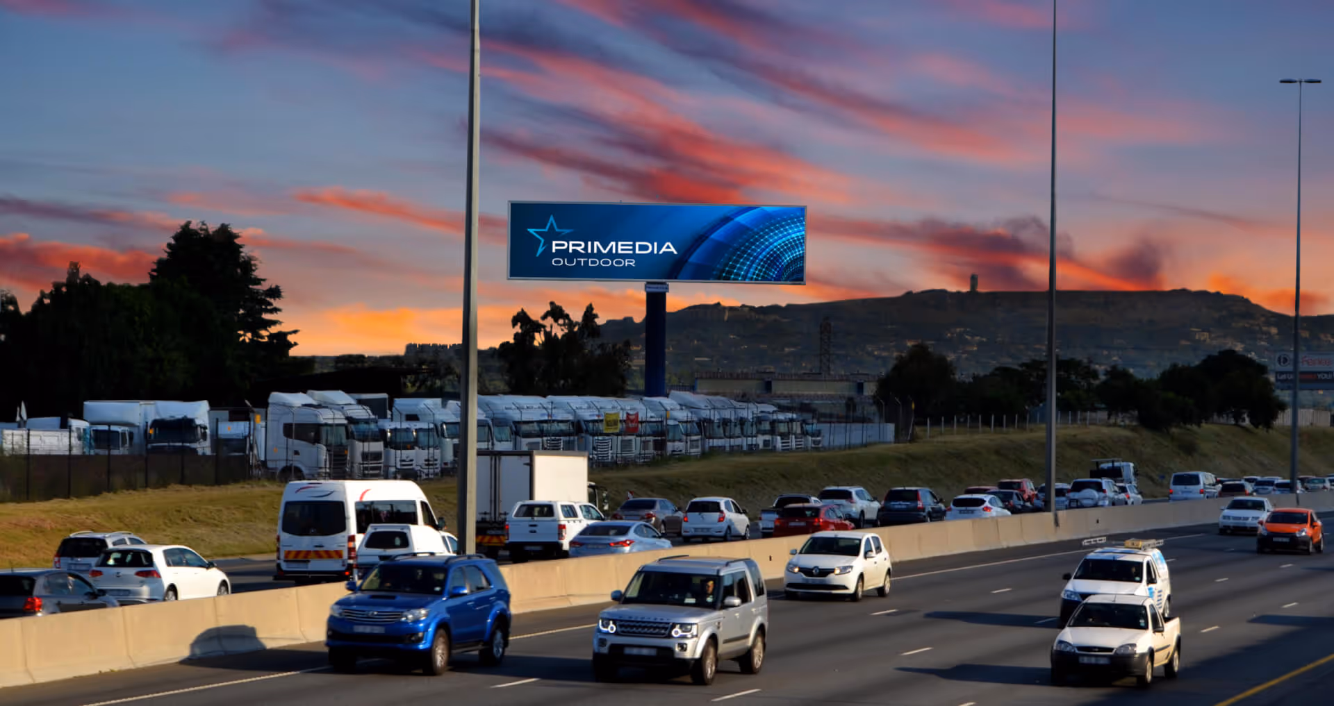 Highway with multiple cars and trucks passing under an electronic billboard at sunset.