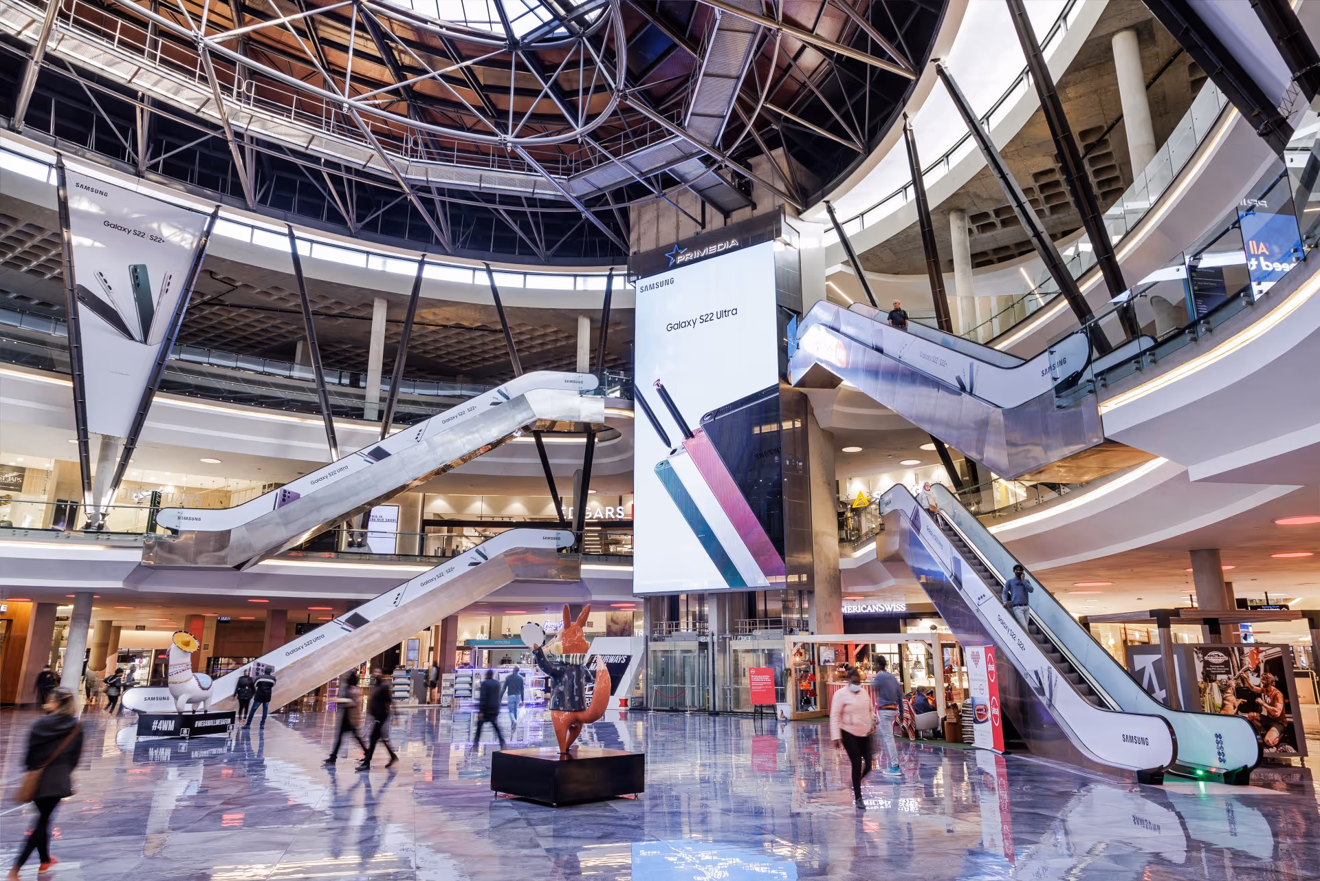 Shopping mall interior with Samsung Galaxy S22 Ultra ads on escalators and a large digital display.