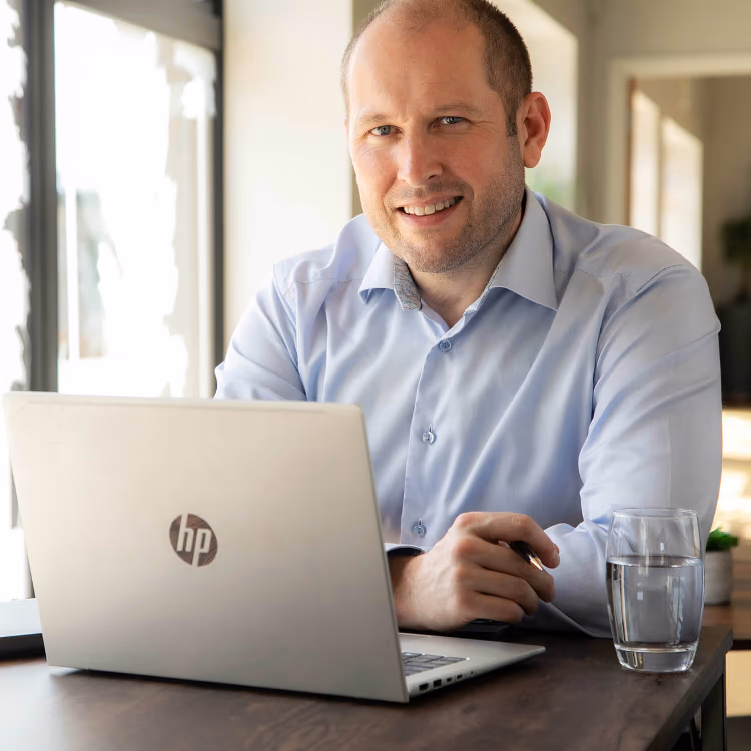 Man in light blue shirt smiling, sitting at a table with a laptop and a glass of water.