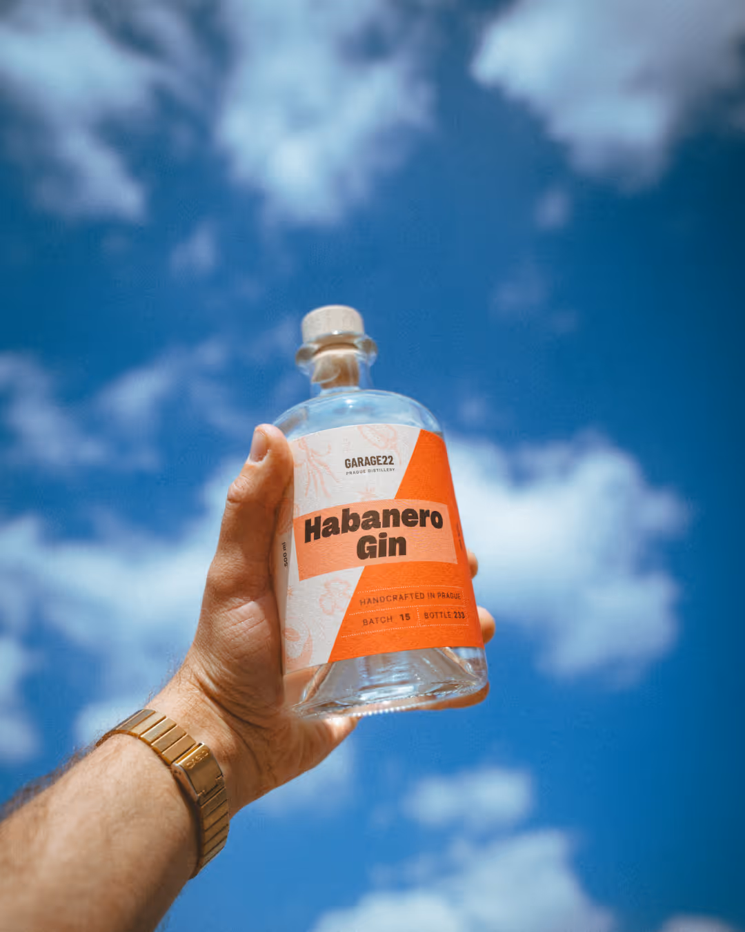 Ruka držící láhev habanero ginu značky Garage22 proti modré obloze s mraky.