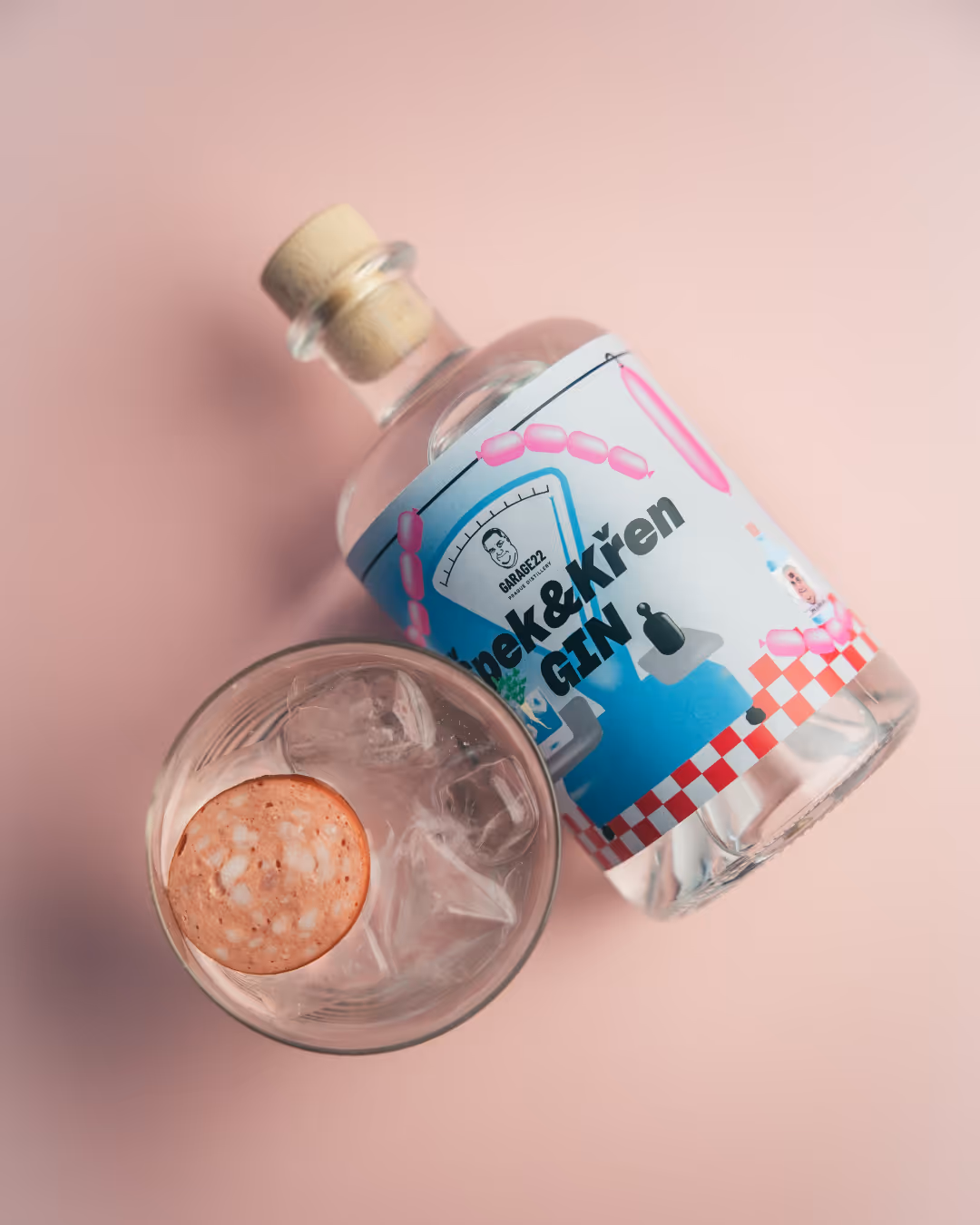 Lahvička s nápisem Spek & Křen GIN vedle sklenice s ledem a plátkem salámu na růžovém pozadí.