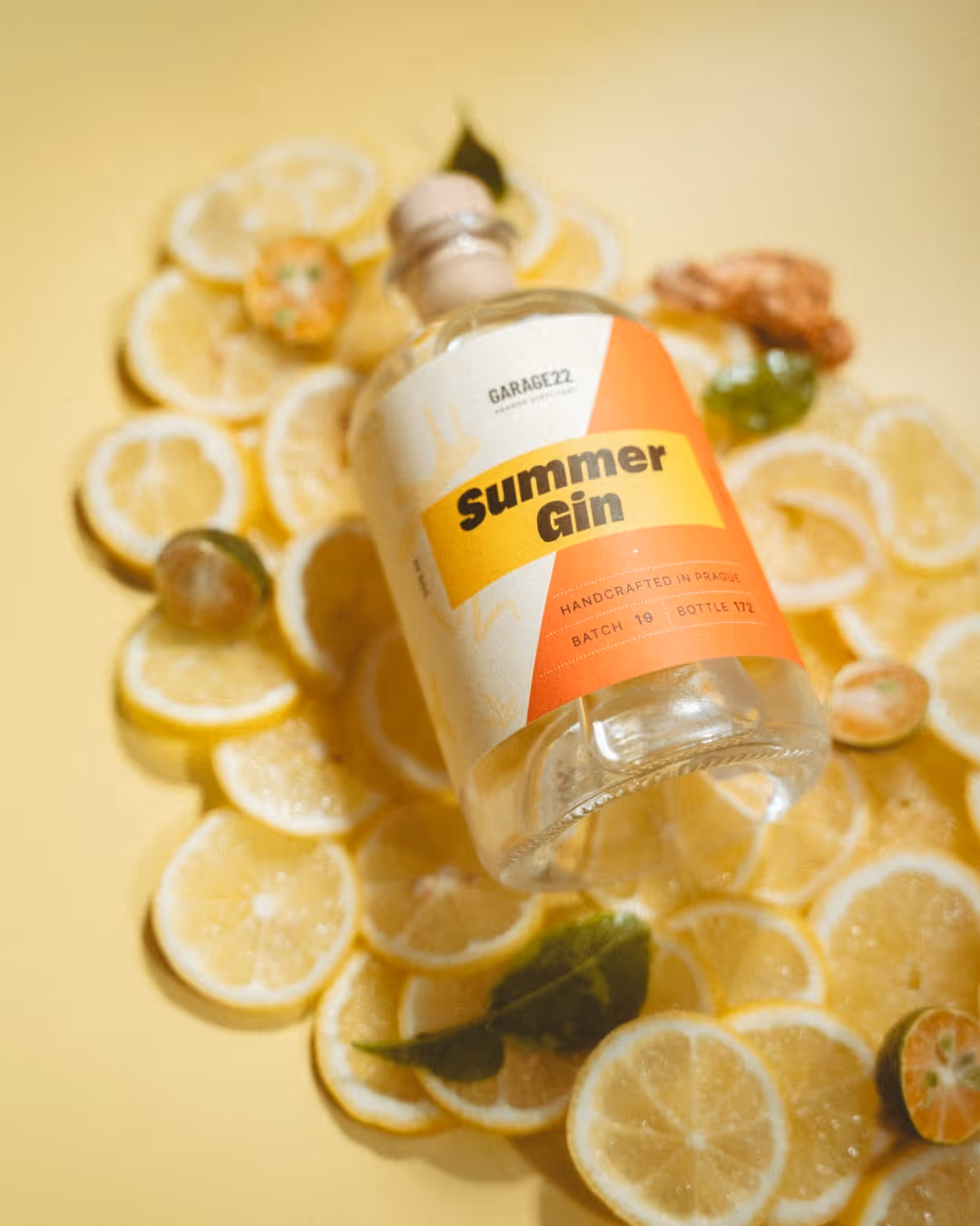 Láhev letního ginu GARAGE22 položena na svazek plátků citronu a zelených listů na žlutém pozadí.
