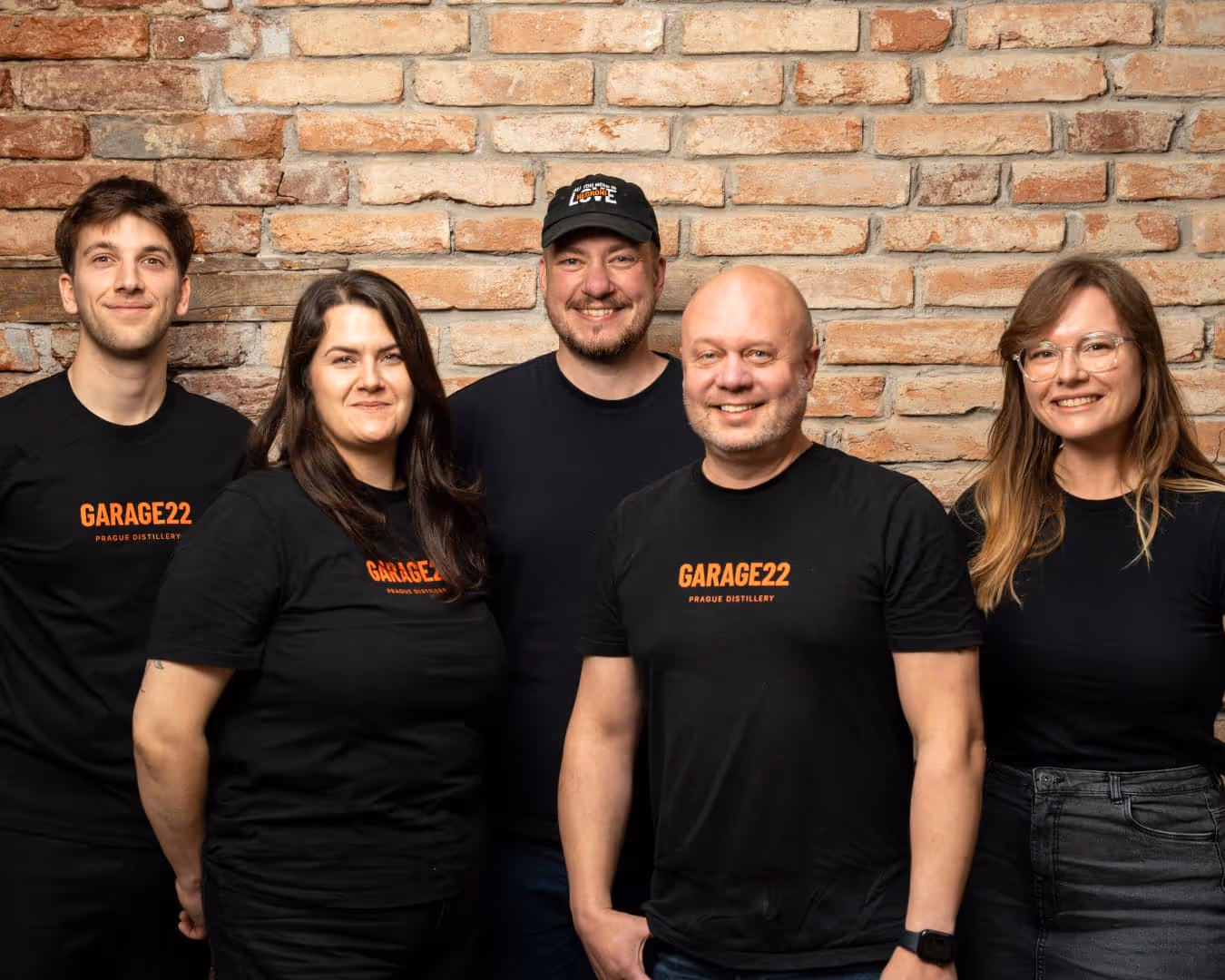 Pět členů týmu stojí před cihlovou zdí, tři z nich mají černá trička s nápisem GARAGE22 Prague Distillery.
