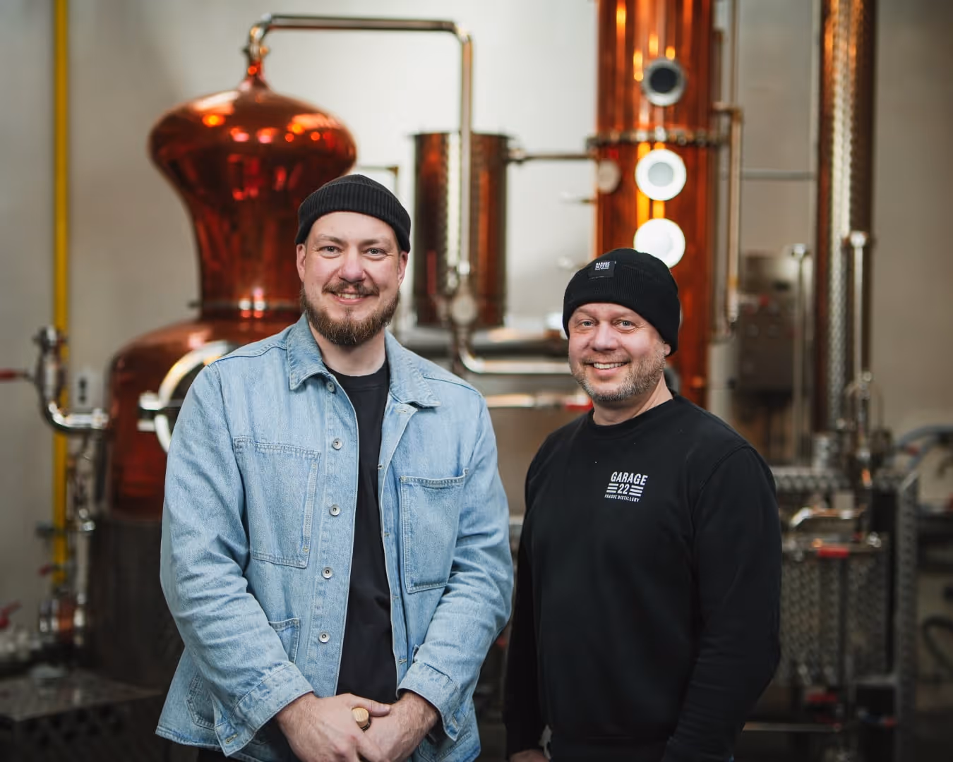 Dva muži v garážové palírně stojí před měděným destilačním zařízením, oba mají na hlavě čepice a usmívají se.