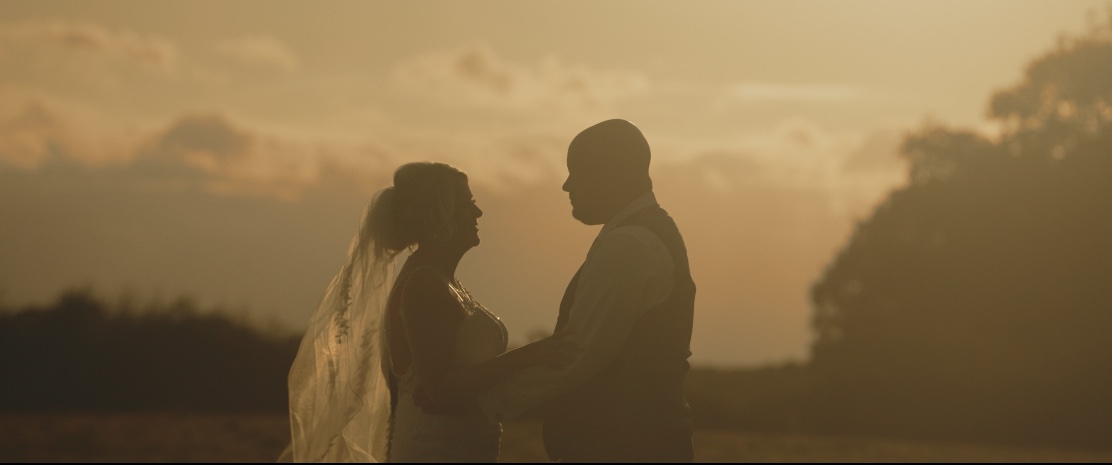 Silhouetted bride and groom facing each other and holding hands outdoors at sunset.