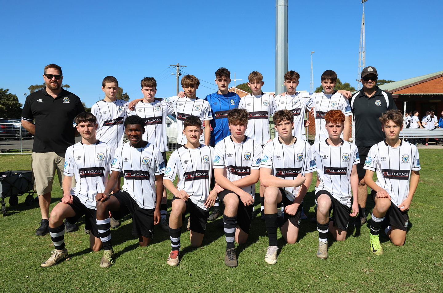 Youth soccer team in white and black kits posing outdoors on grass with two adult coaches on either side under a clear blue sky.