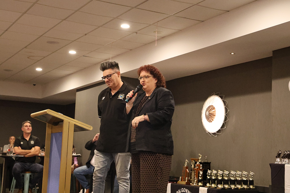 A woman speaking into a microphone and a man standing beside her at a podium in a room with trophies on a table.