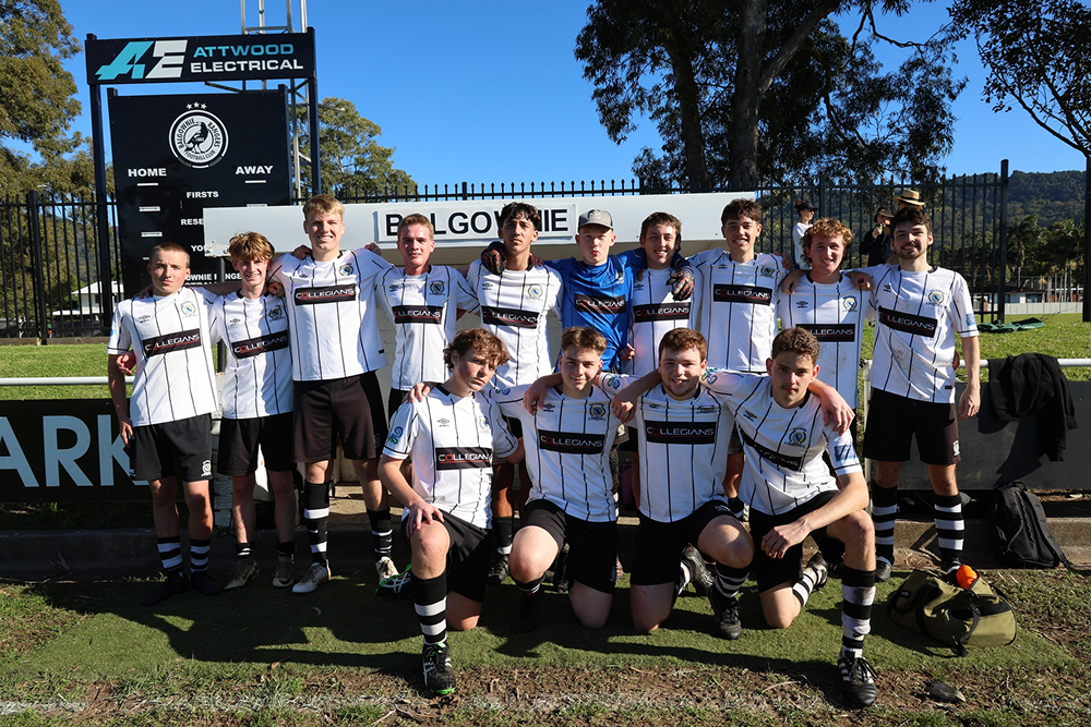 Male soccer team in white and black striped uniforms posing together on a grassy field with a scoreboard and trees in the background.
