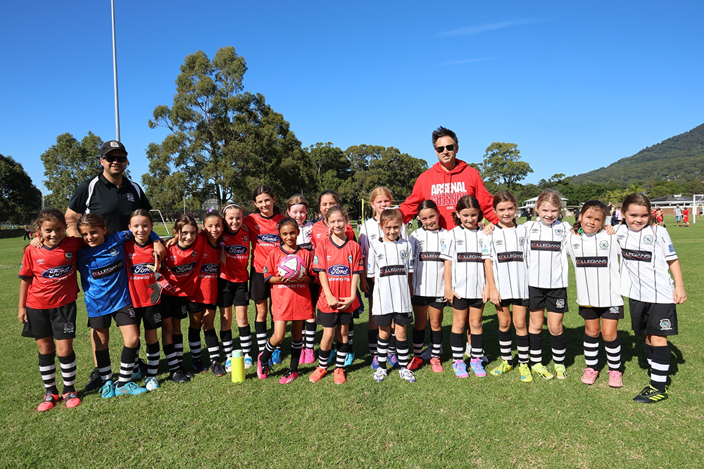 Girls youth soccer teams in red and white uniforms posing together on a sunny grassy field with two coaches behind them.