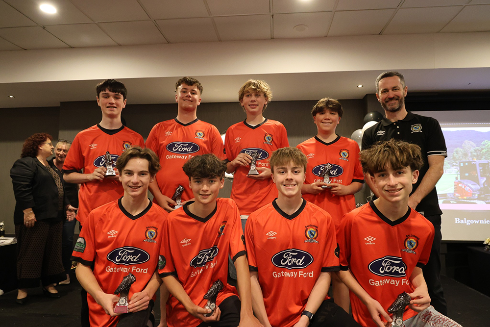 Youth soccer team in orange Ford Gateway Ford jerseys holding individual player trophies, standing and kneeling indoors with a coach.