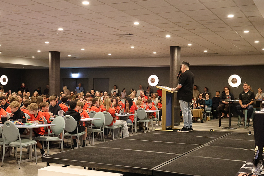 Man speaking into a microphone at a podium in front of a seated audience, including children in orange shirts, in a large conference room.