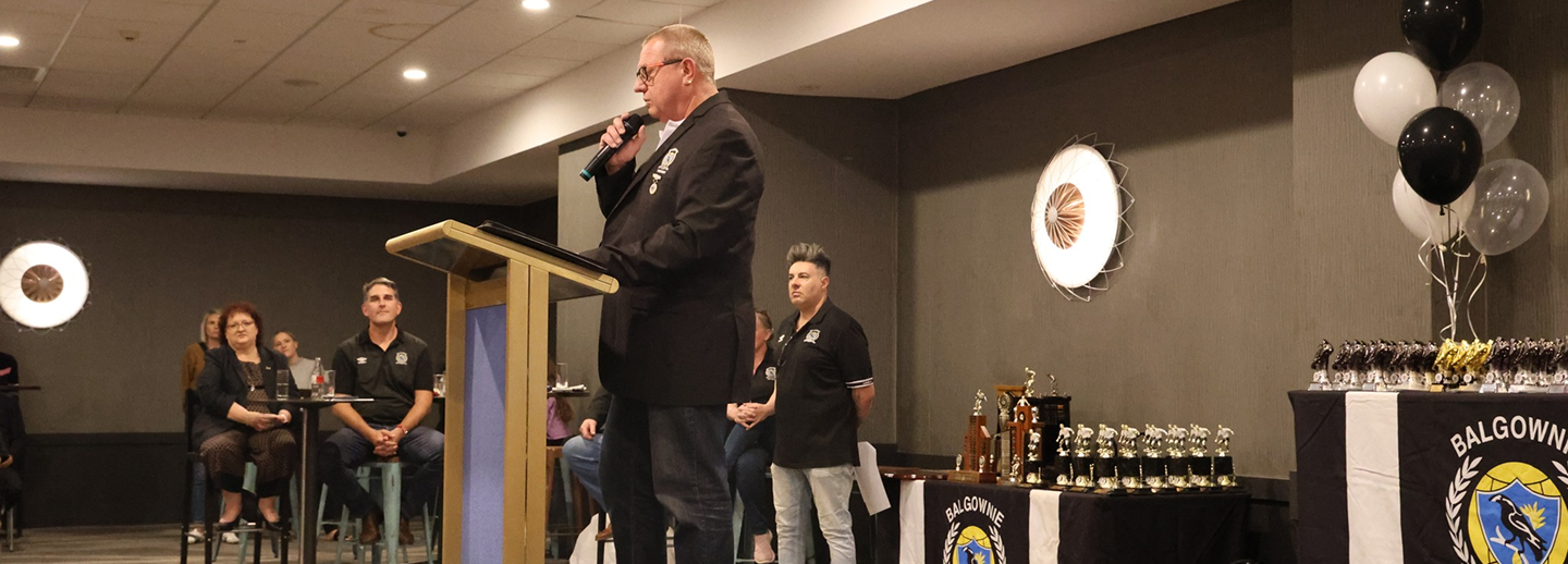 Man in a black blazer speaking into a microphone at a podium during an indoor event with trophies and balloons on tables nearby.