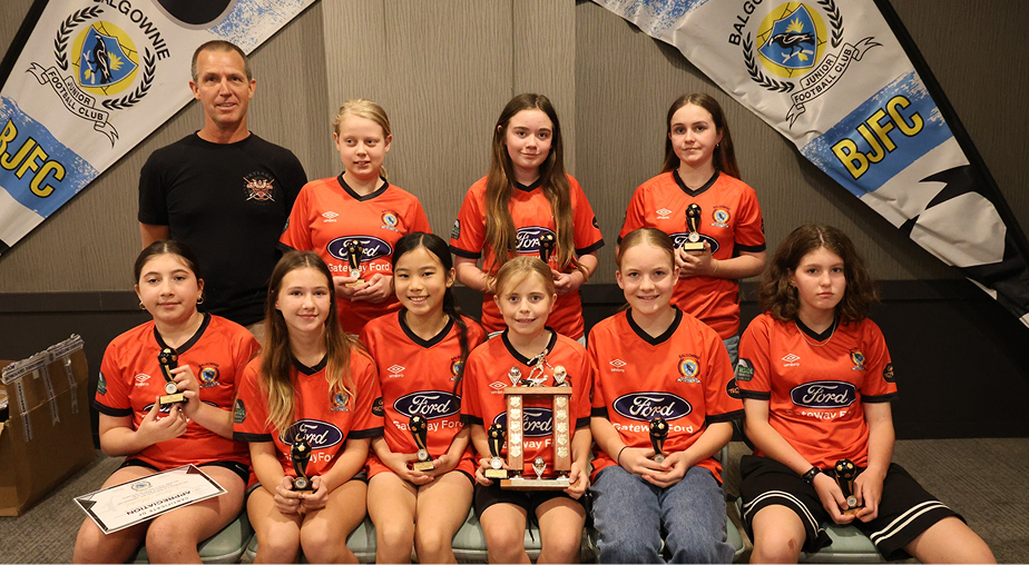 Youth soccer team wearing orange jerseys holding trophies and a large shield, posing indoors with two BJFC banners in the background.
