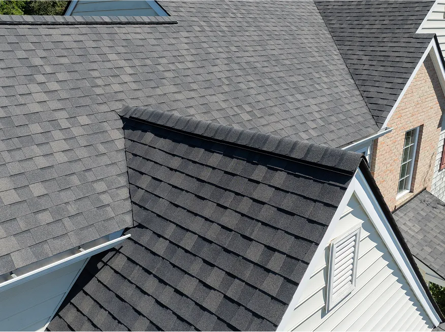 A close-up view of a house's roof with dark gray asphalt shingles and white siding, showing multiple roof angles and a vent.