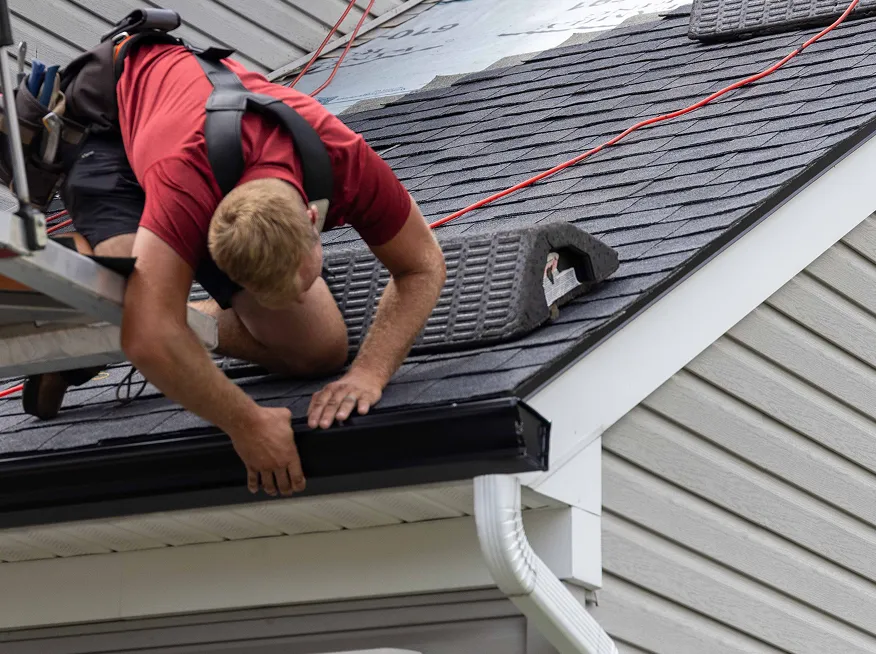 A roofer on a ladder installs a black rain gutter on the edge of a house roof, with his hands securing the gutter in place.