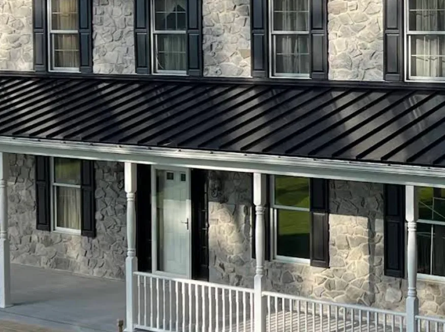 A stone house with black metal roofing, black shutters, multiple windows, a white front door, and a white-railed porch.