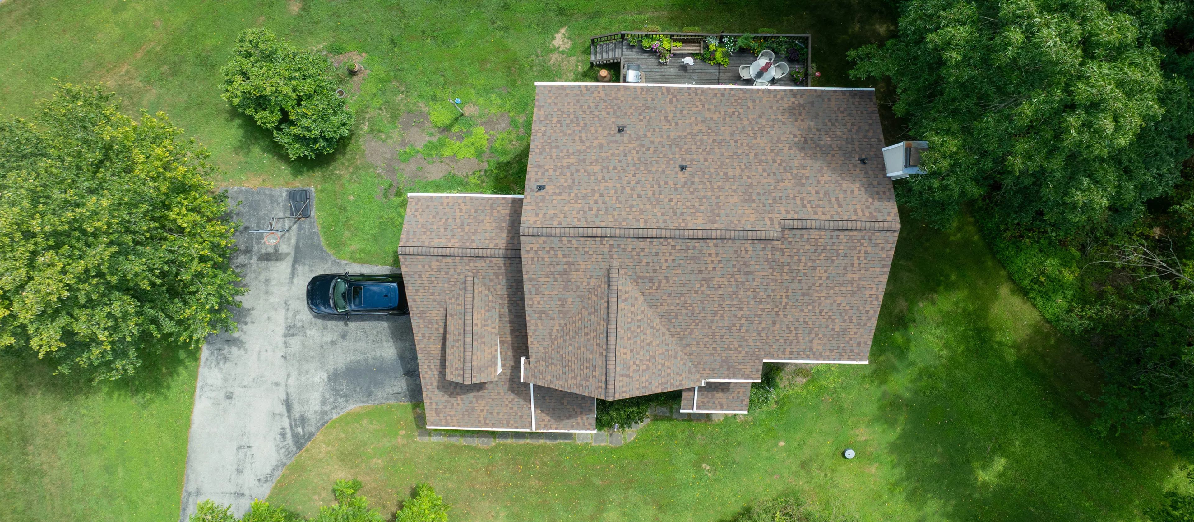 Aerial view of a house with a brown roof, driveway with a parked car, deck with patio furniture, and surrounding green trees and lawn.