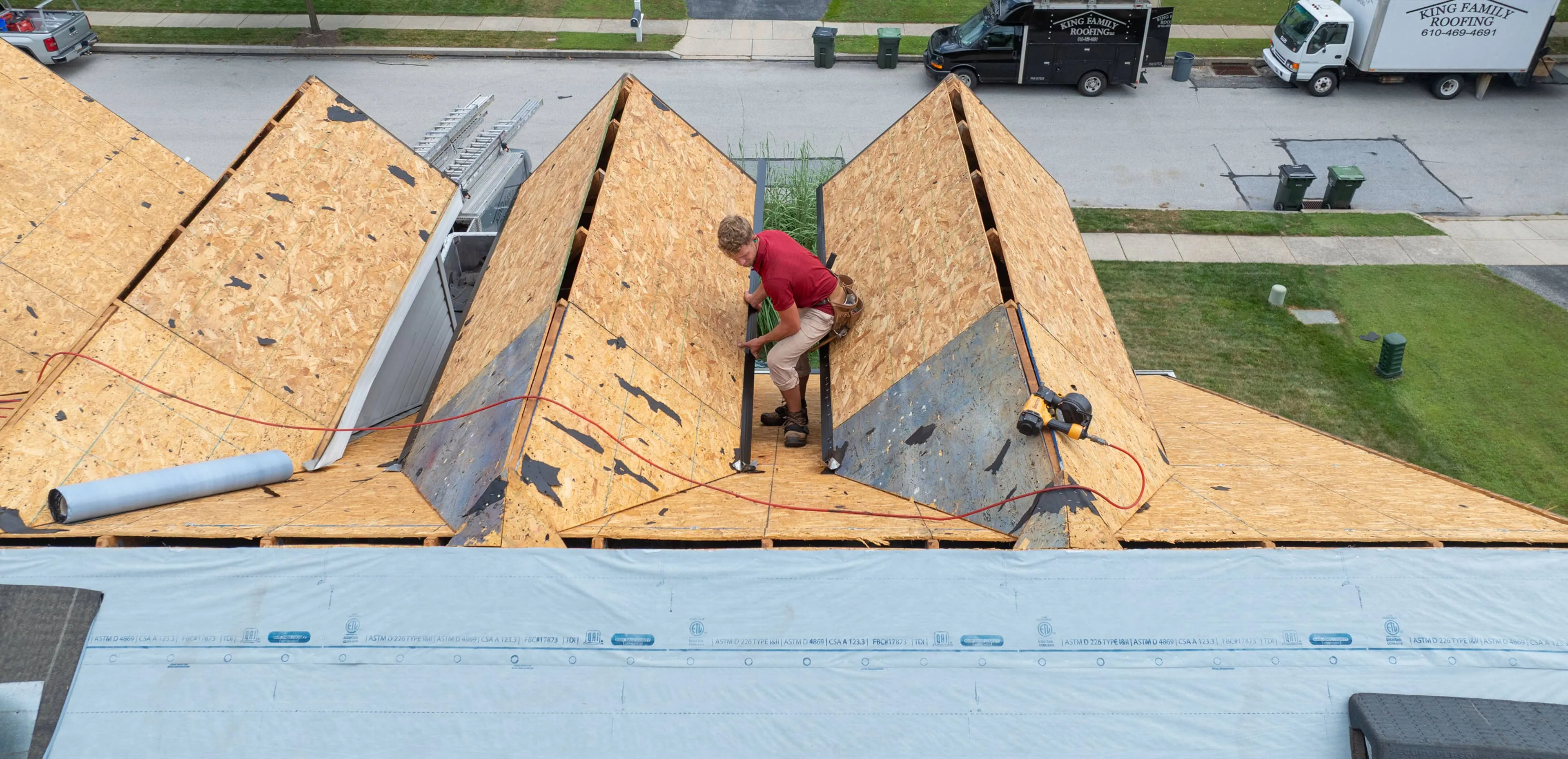 A worker in a red shirt installs plywood roof sheathing on a house under construction.