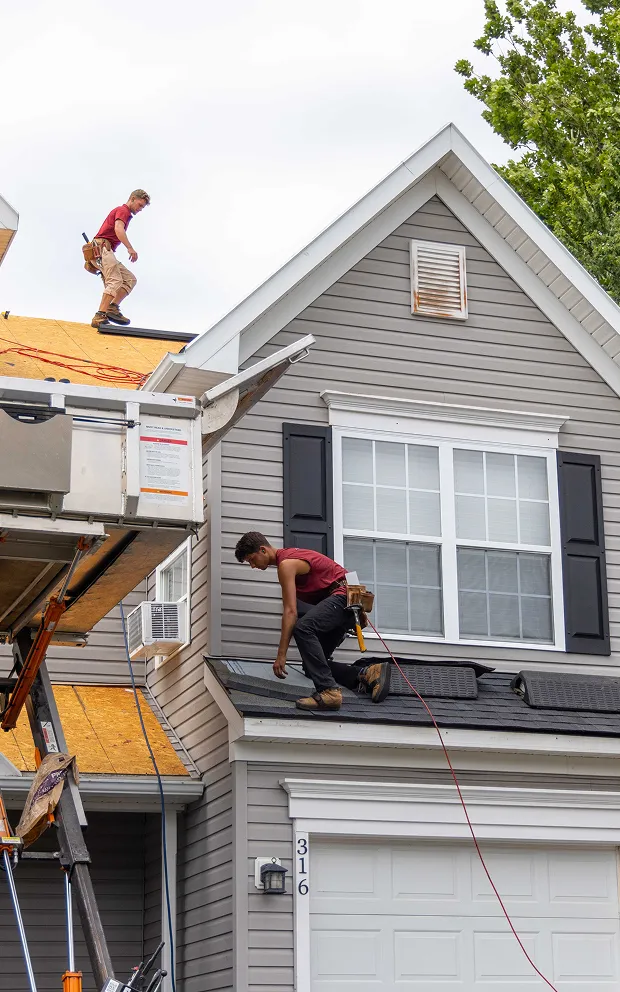 Two roofers install shingles on the roof of a gray house with white trim, using safety harnesses and tools.