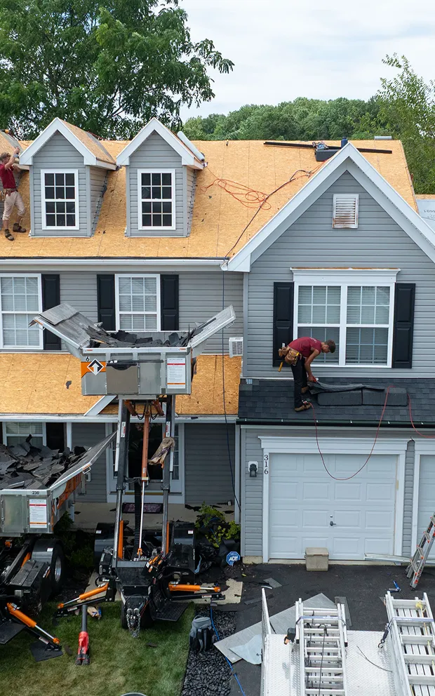 Workers are installing new plywood roof decking on a two-story gray house.