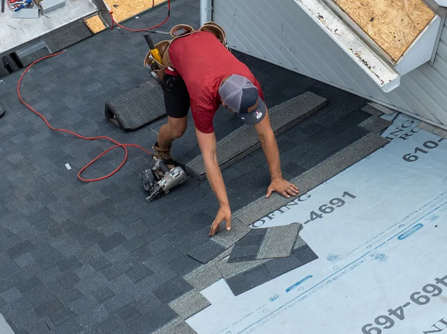 A worker in a red shirt is installing asphalt shingles on a house roof using a nail gun.
