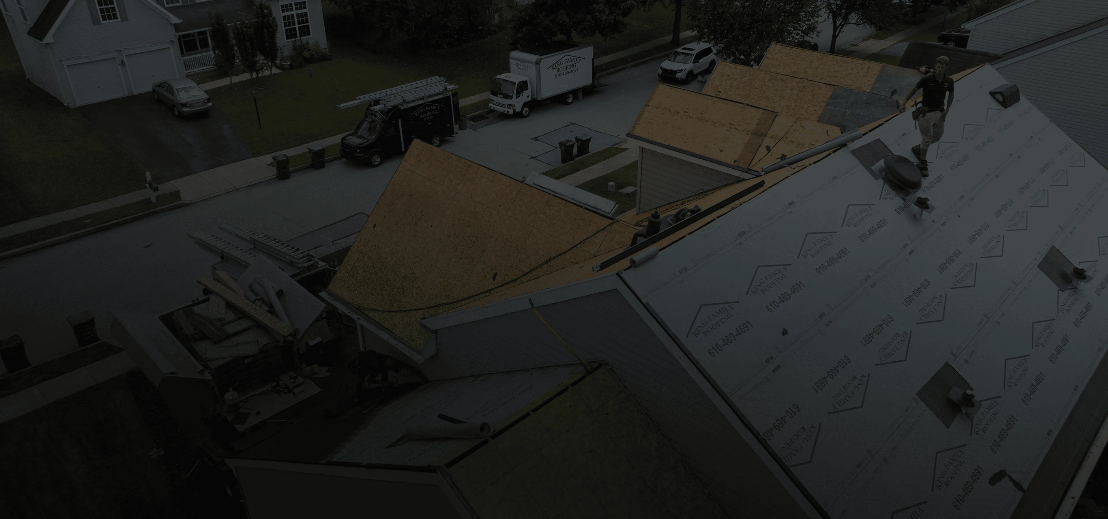 Workers install new roofing materials on several adjacent houses in a suburban neighborhood, with construction vehicles and equipment visible on the street below.