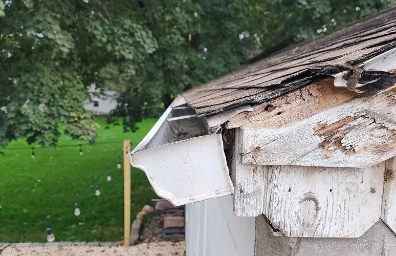 A house roof corner with damaged wood and shingles, showing a broken gutter hanging loosely.
