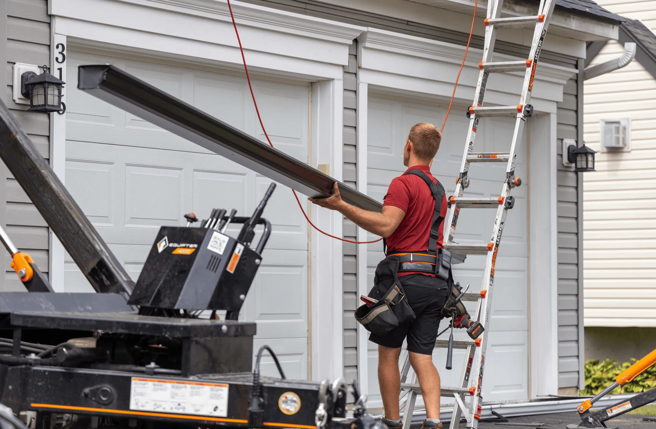 A roofer wearing a tool belt carries a long metal gutter toward a two-car garage, with equipment and a ladder nearby.