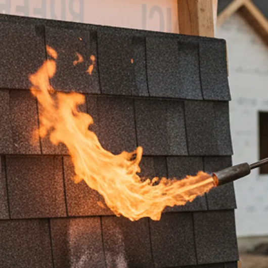 A torch flame is being applied to black roof shingles, demonstrating a fire resistance test on roofing material.