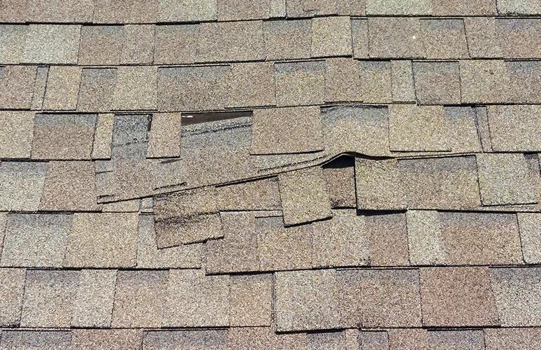 Damaged asphalt roof shingles with several pieces lifted and misaligned, exposing the underlying roof structure.