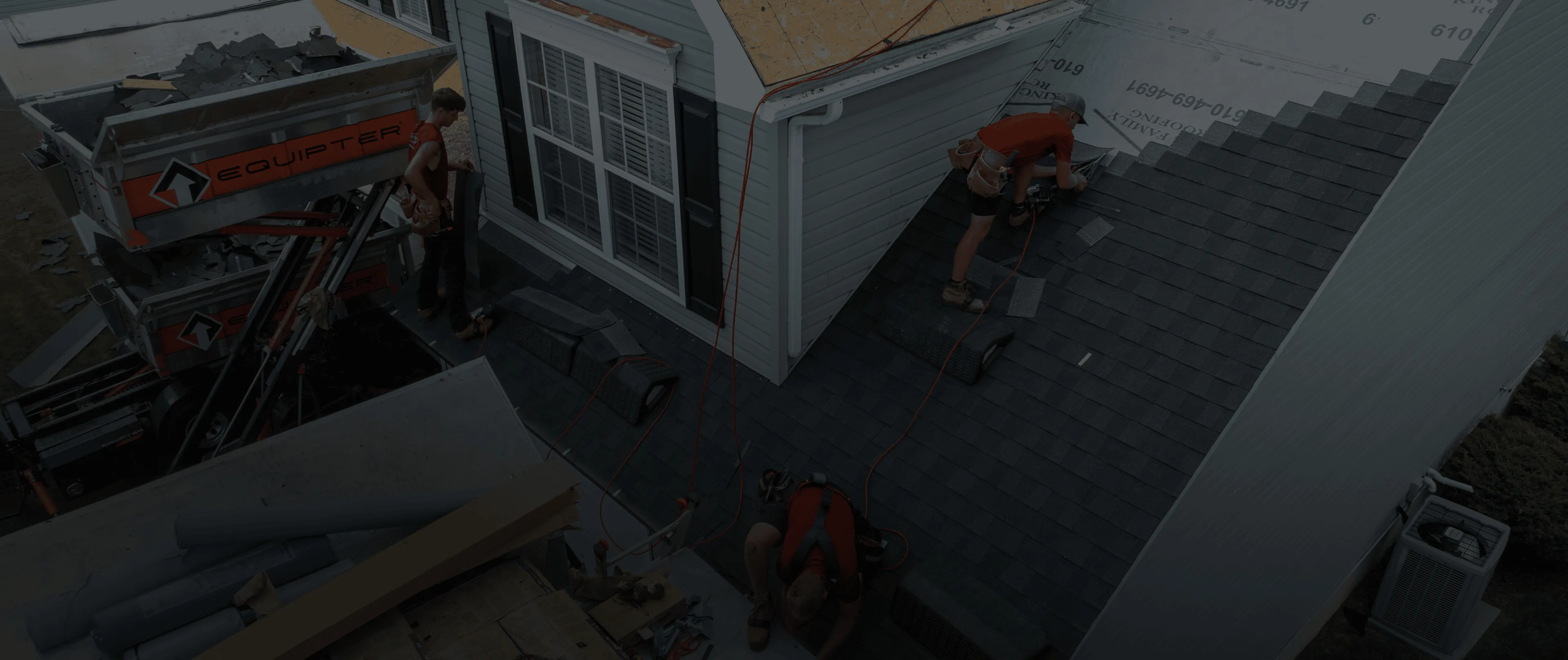 Three roofers install shingles on a house roof; one stands near equipment, while two others work on the sloped roof surface.