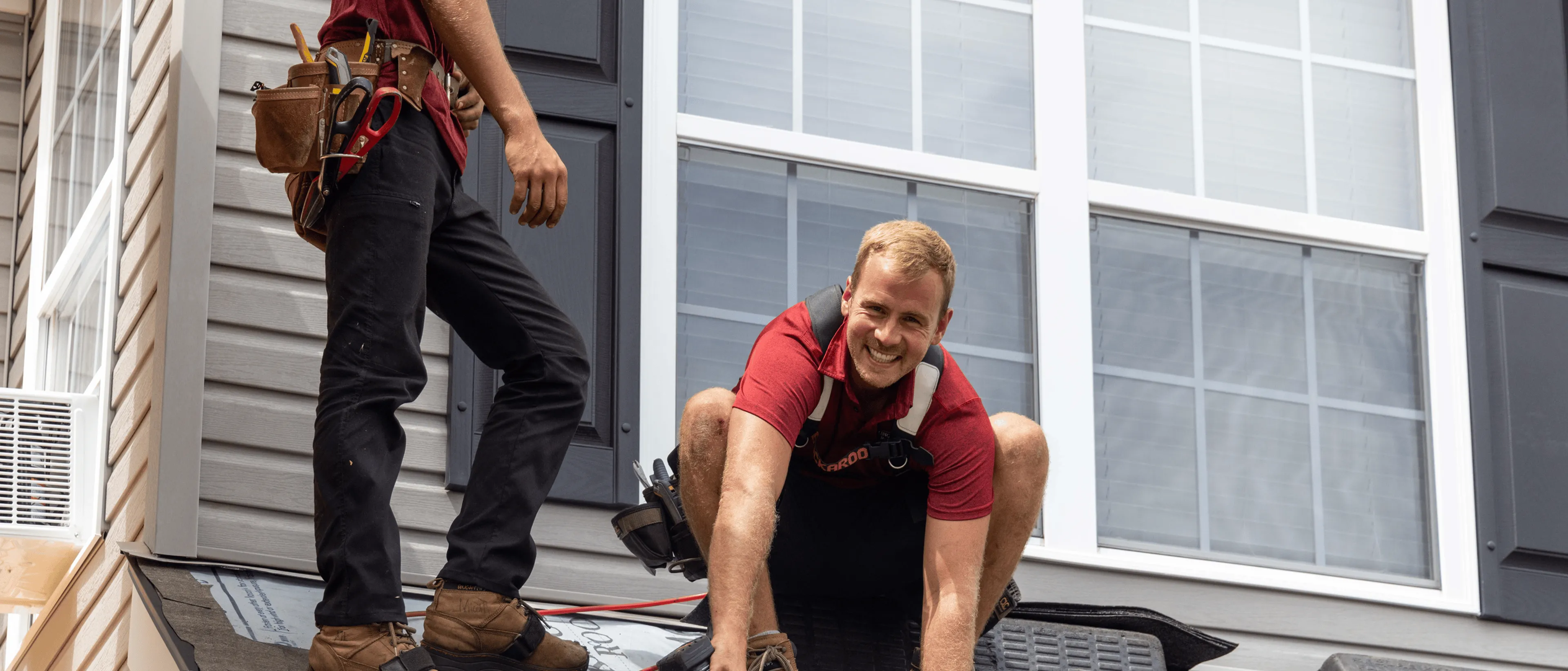 Two workers in red shirts and tool belts work on the roof of a house in front of large windows.