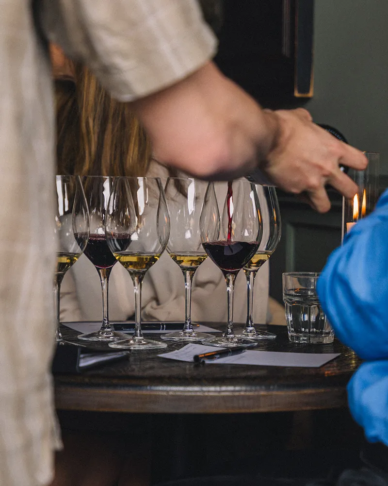 Person som häller upp rött vin i ett av flera vinglas på ett bord under en vinprovning.