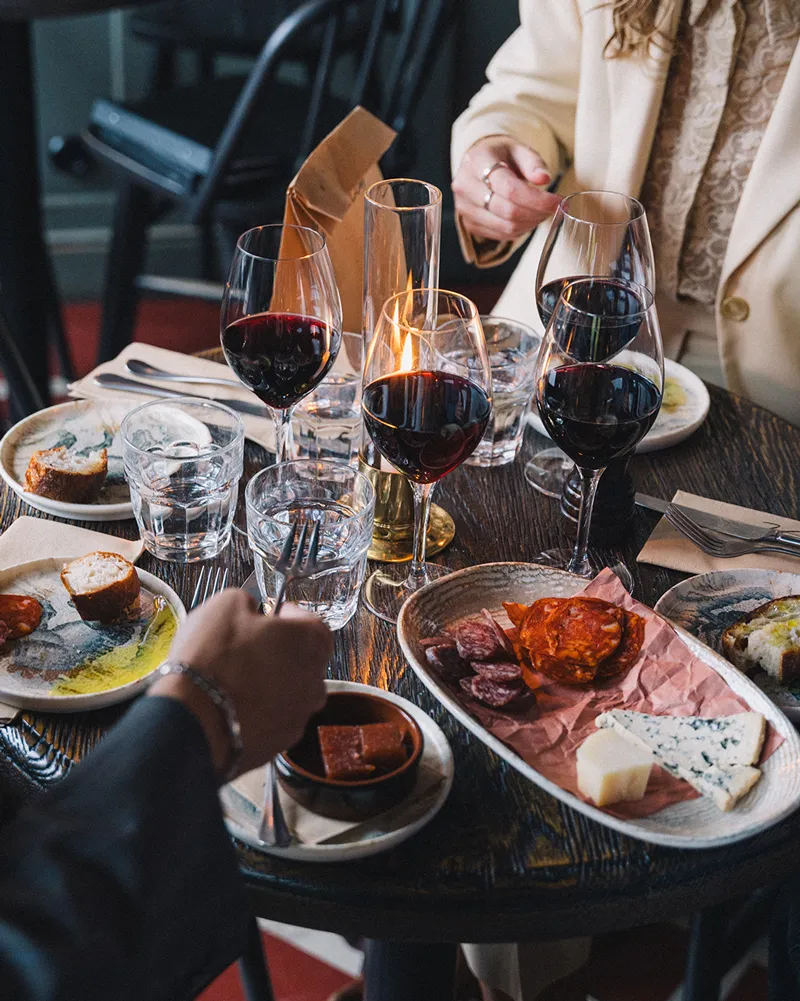 Bord med glas rött vin, tallrikar med olika ostar och charkuterier, samt personer som äter.