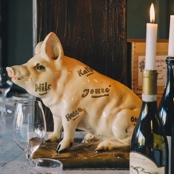 Keramisk grisfigur med namn skrivna på kroppen, placerad på ett bord med vinflaskor, ett ljus och glas.