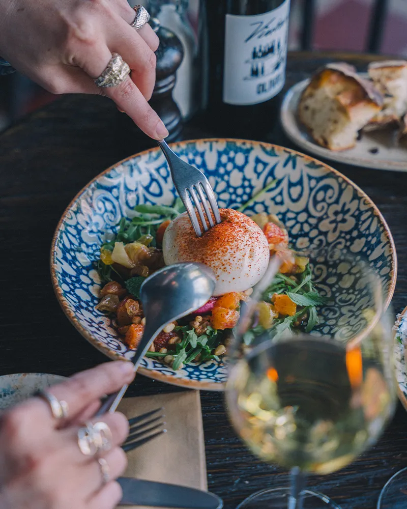 Händer som använder gaffel och sked för att skära i en rund ost på en bladig sallad, serverad i en dekorativ blåvit skål, med bröd och ett glas vitt vin på bordet.