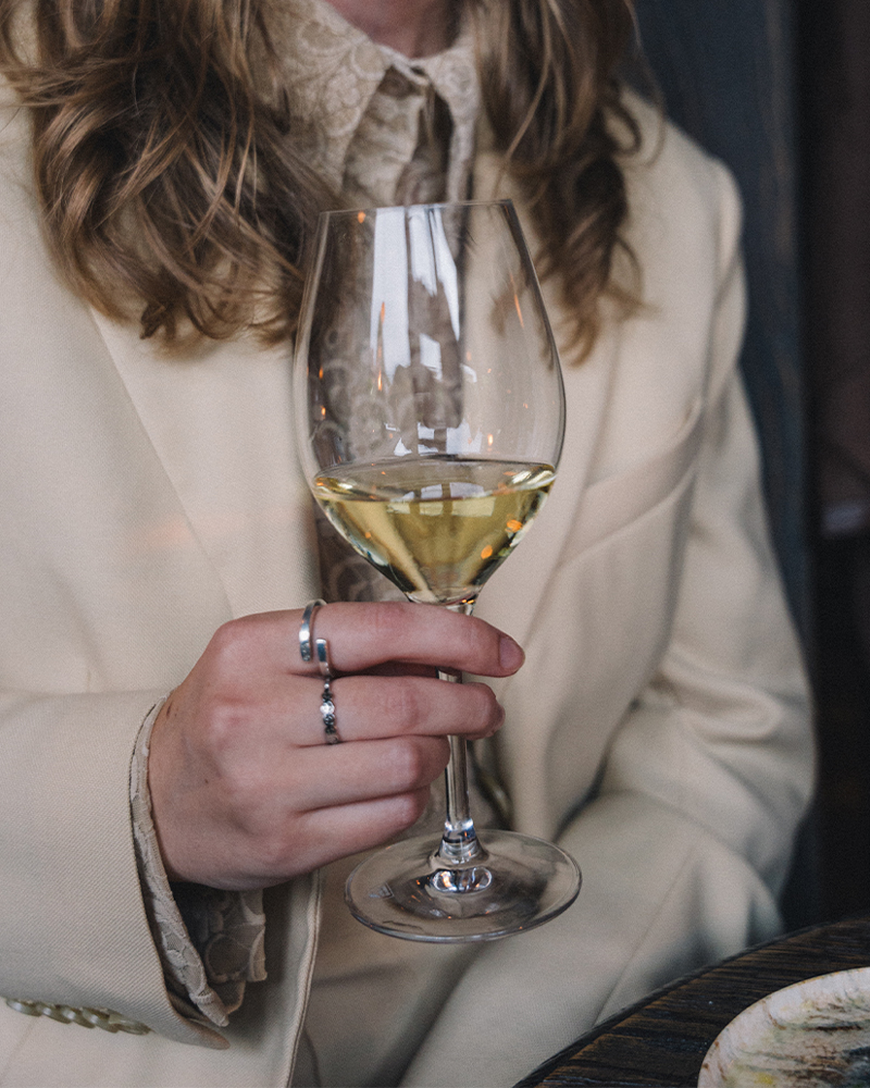 Person i krämfärgad blazer som håller ett glas vitt vin vid ett bord.