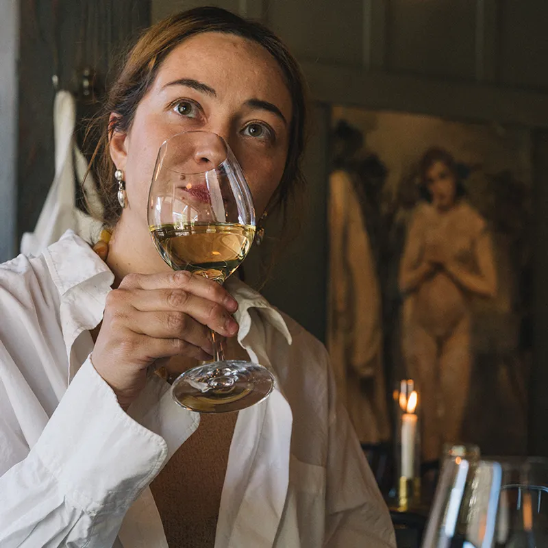 Kvinna i vit skjorta som håller ett glas vitt vin nära ansiktet, med en mjukt upplyst bakgrund med en målning och tända ljus.