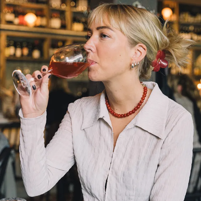 Blond kvinna med rött pärlhalsband som dricker rött vin ur ett vinglas i en mysig vinbar.